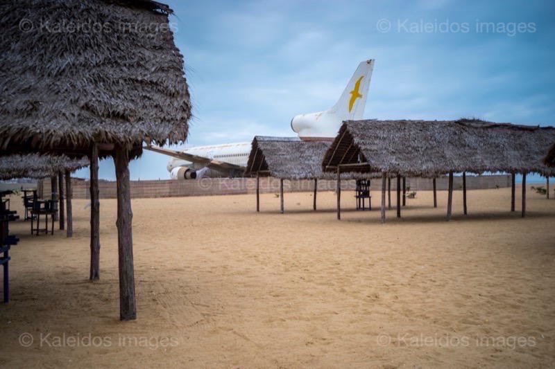 Barakah, L1011 TriStar, Lockheed L1011, abandoned TriStar, aircraft wreck, grounded aircraft, white fuselage, derelict cockpit, empty cabin, Cotonou beach, Cadjéhoun beach, Cotonou Benin, West Africa, Benin airport, Atlantic coastline, coastal Benin, corroded wreck, marine rust, deterioration, salt corrosion, decay, abandonment, dereliction, dilapidation, environmental erosion, aviation heritage, industrial archaeology, failed tourism project, open-air museum, urban art, colonial relic, Benin history, alternative tourism, architectural curiosity, aviation history, hajj pilgrimage, Air Rum airline, defunct carrier, humanitarian transport, international migration, historical traces, industrial heritage, stranded aircraft, tourist attraction, urban exploration, ruin photography, beach adventure, Beninese leisure, amusement park, immersive experience, urban safari, overcast sky, sunset beach, tropical storm, coastal erosion, sea salt, Atlantic Ocean, white sand, coastal vegetation, gleaming white, nature-machine contrast, aerospace geometry, outstretched wings, empty windows, cabin stairway, cockpit instruments, time displacement, obsolescence, suspended time, liminality, aborted transformation, urban memory, unfinished dream, nature-industry symbiosis, tourism ghost, frozen history
