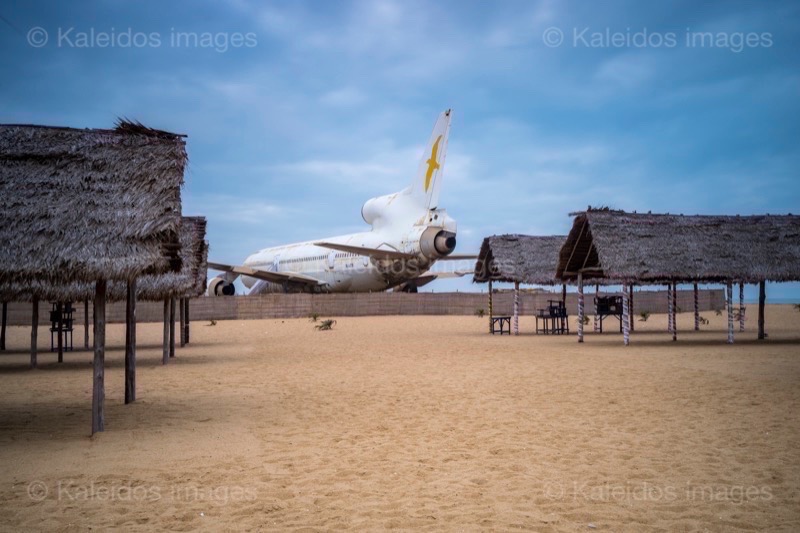 Barakah, L1011 TriStar, Lockheed L1011, abandoned TriStar, aircraft wreck, grounded aircraft, white fuselage, derelict cockpit, empty cabin, Cotonou beach, Cadjéhoun beach, Cotonou Benin, West Africa, Benin airport, Atlantic coastline, coastal Benin, corroded wreck, marine rust, deterioration, salt corrosion, decay, abandonment, dereliction, dilapidation, environmental erosion, aviation heritage, industrial archaeology, failed tourism project, open-air museum, urban art, colonial relic, Benin history, alternative tourism, architectural curiosity, aviation history, hajj pilgrimage, Air Rum airline, defunct carrier, humanitarian transport, international migration, historical traces, industrial heritage, stranded aircraft, tourist attraction, urban exploration, ruin photography, beach adventure, Beninese leisure, amusement park, immersive experience, urban safari, overcast sky, sunset beach, tropical storm, coastal erosion, sea salt, Atlantic Ocean, white sand, coastal vegetation, gleaming white, nature-machine contrast, aerospace geometry, outstretched wings, empty windows, cabin stairway, cockpit instruments, time displacement, obsolescence, suspended time, liminality, aborted transformation, urban memory, unfinished dream, nature-industry symbiosis, tourism ghost, frozen history